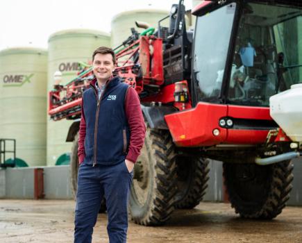 Image of a man with brown hair and navy blue gilet jacket stood in front of a red agricultural crop sprayer.