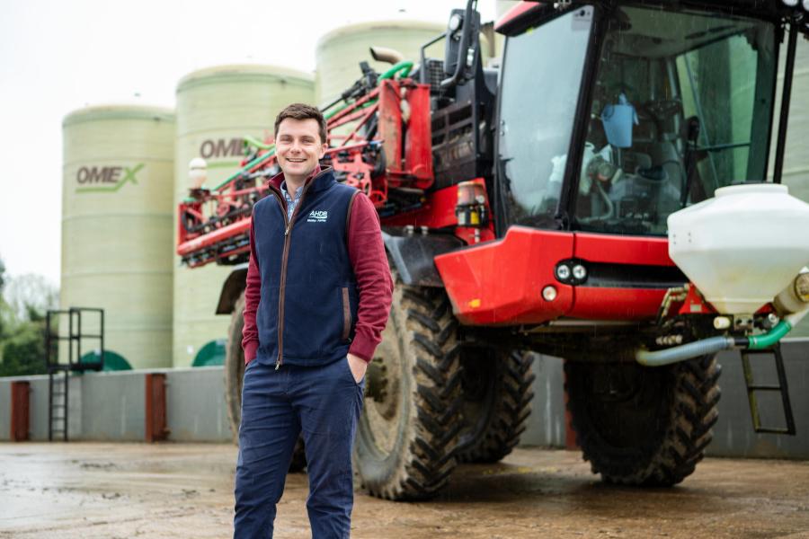 Image of a man with brown hair and navy blue gilet jacket stood in front of a red agricultural crop sprayer.