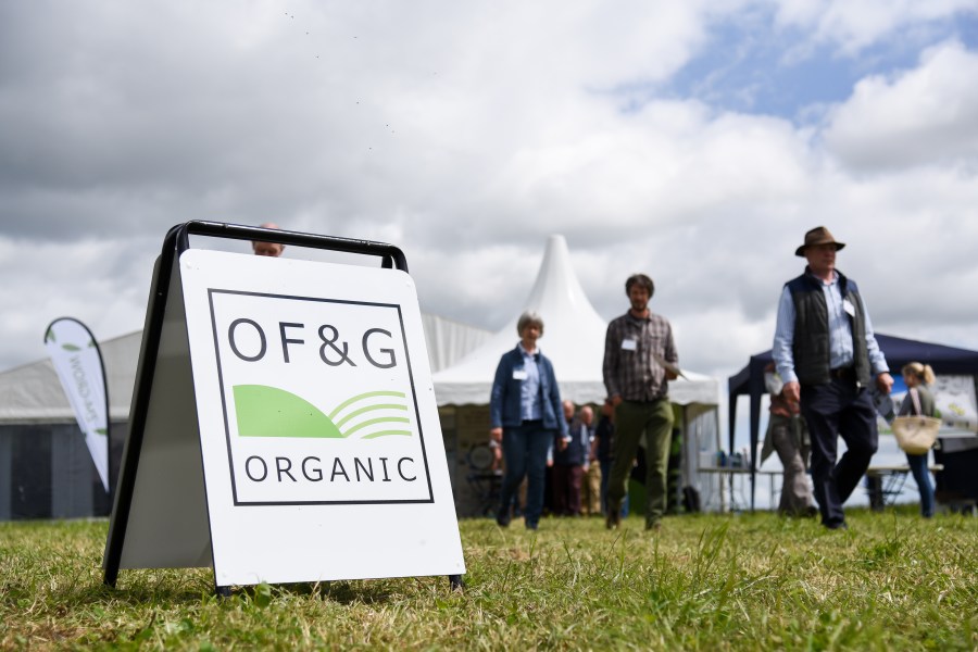 Image of an agricultural event in a field, with a board featuring the OF & G organic logo and people walking to and from a large bell tent.