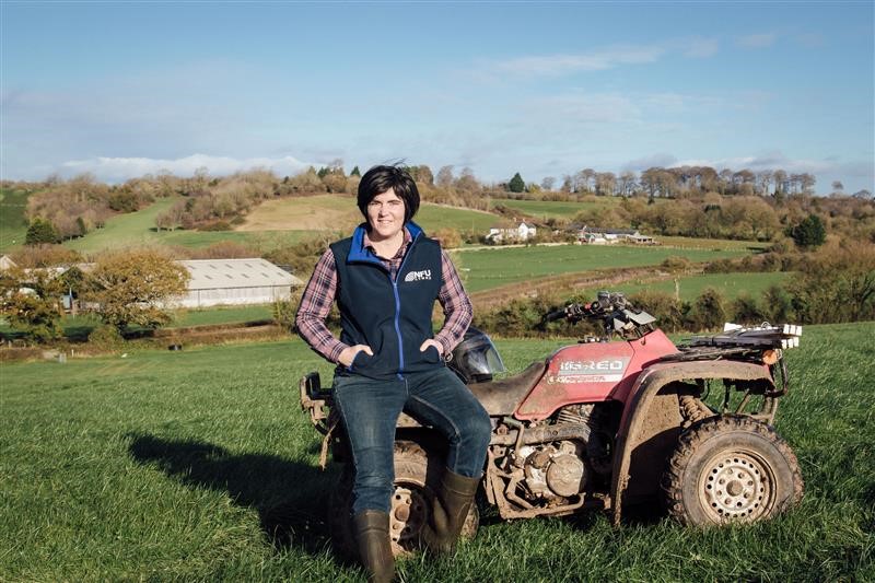 An image of a women with dark hair sat on the front of a quad bike, set against a farming landscape with a blue sky.