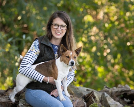 Image of a young lady called Chloe Timberlake who is sat on a stone wall with a small terrier type dog on her knee, against a background of leafy countryside.