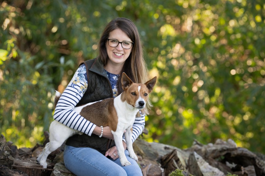 Image of a young lady called Chloe Timberlake who is sat on a stone wall with a small terrier type dog on her knee, against a background of leafy countryside.