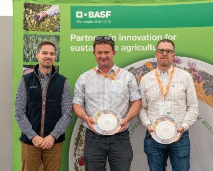 Image of three men standing together in front of a large, wide green marketing banner, two of whom are holding commemorative plates.