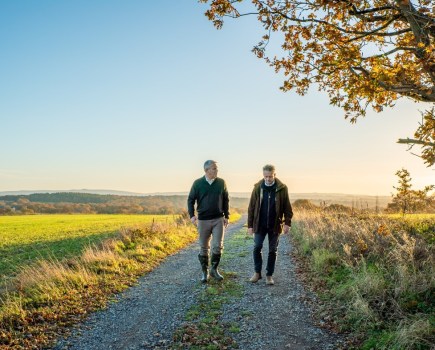 Image of two people walking in parallel along a farm track or similar, surrounded by countryside and backed by a blue sky.