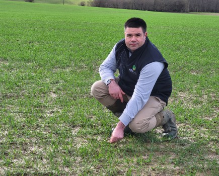 An image of a man with short dark hair crouching in a crop field, wearing a gilet.