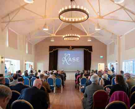 A image of a large hall filled with delegates on seats facing a stage; the stage has a screen with the RASE logo on it.