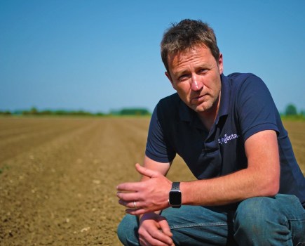 An image of a white man kneeling in a potato field, wearing a blue t-shirt.